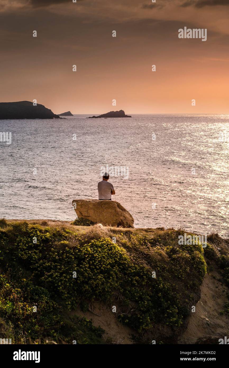 A man sitting alone on a rock on the coast overlooking Fistral Bay in ...