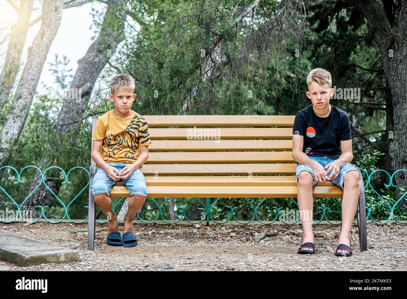 Siblings sitting on wooden bench look with upset and disappointed expressions. Brothers sit on different sides of bench in silence after argument Stock Photo