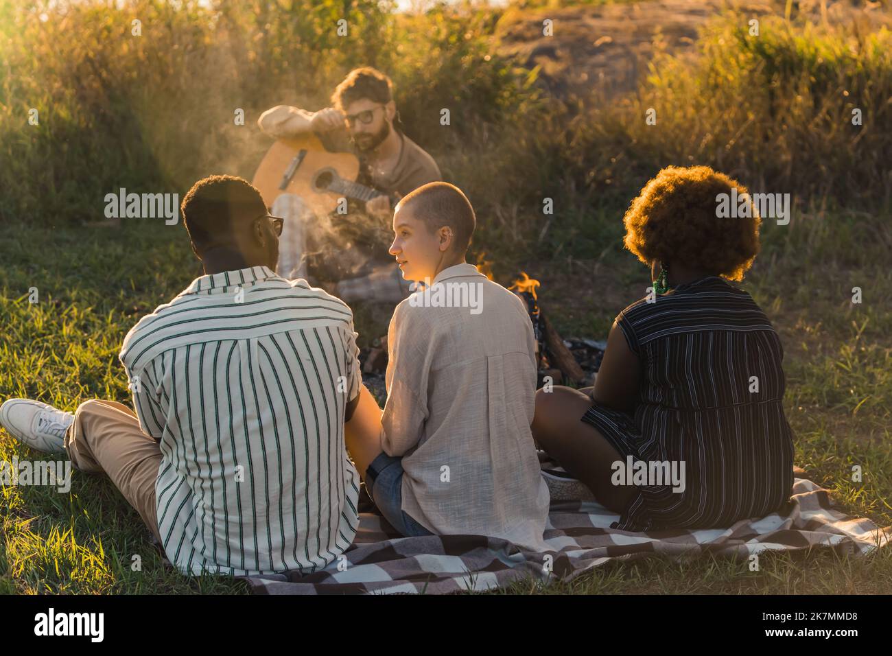 Three people - caucasian queer person, black woman with afro hairstyle, black man in stripped shirt - sitting on checked picnic blanket placed on the ground and listening to their caucasian male friend playing the acoustic guitar. High quality photo Stock Photo