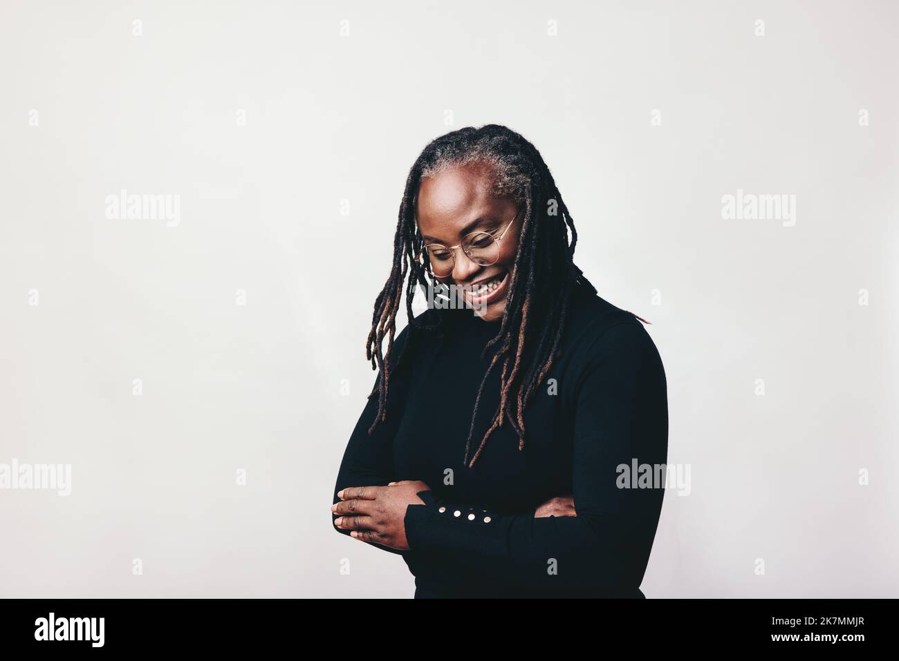 Happy woman with dreadlocks smiling cheerfully while wearing eyeglasses. Professional mature woman standing against a grey background with her arms cr Stock Photo