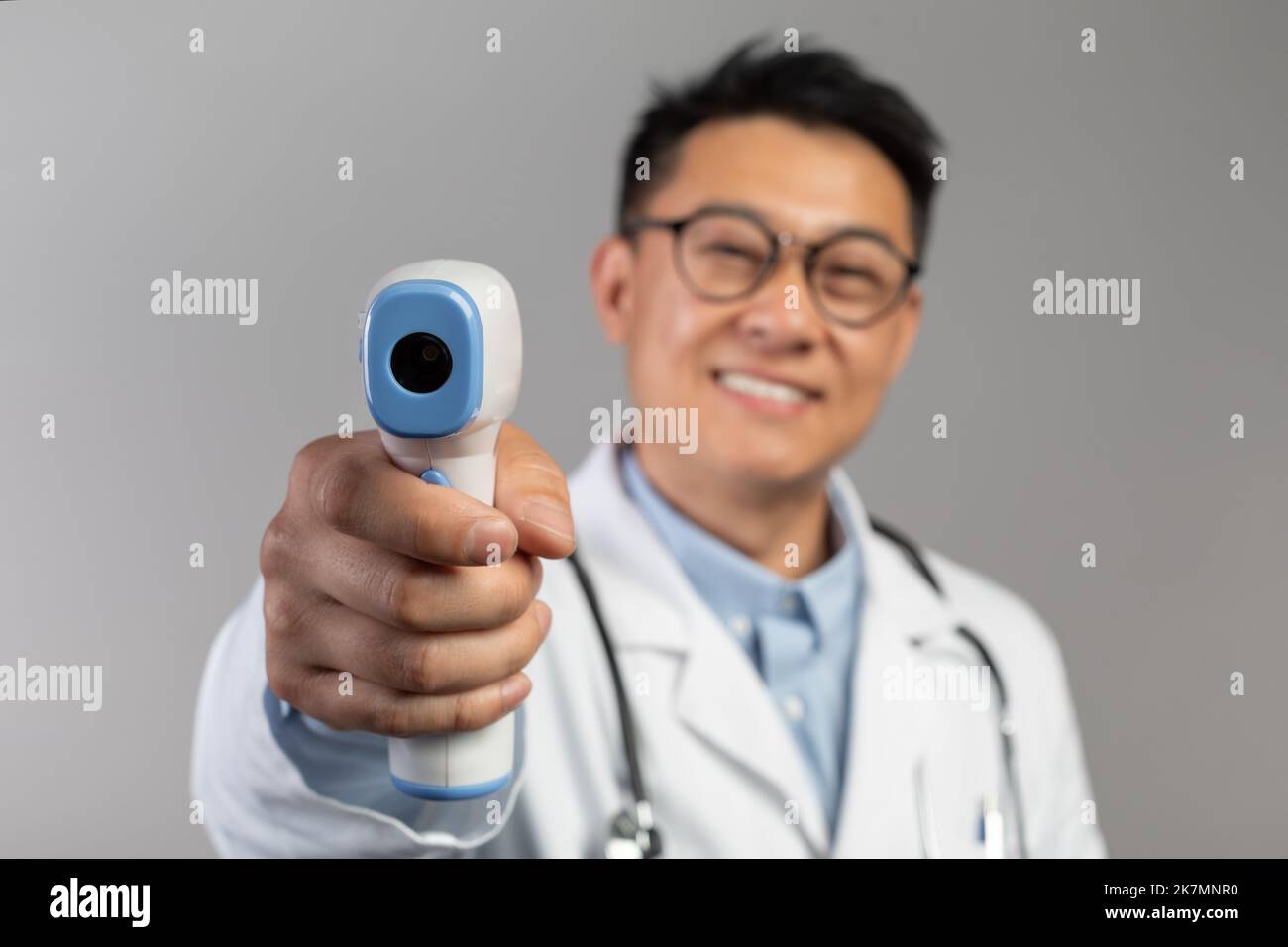 Cheerful middle aged chinese male doctor in white coat, glasses measures temperature with contactless thermometer Stock Photo