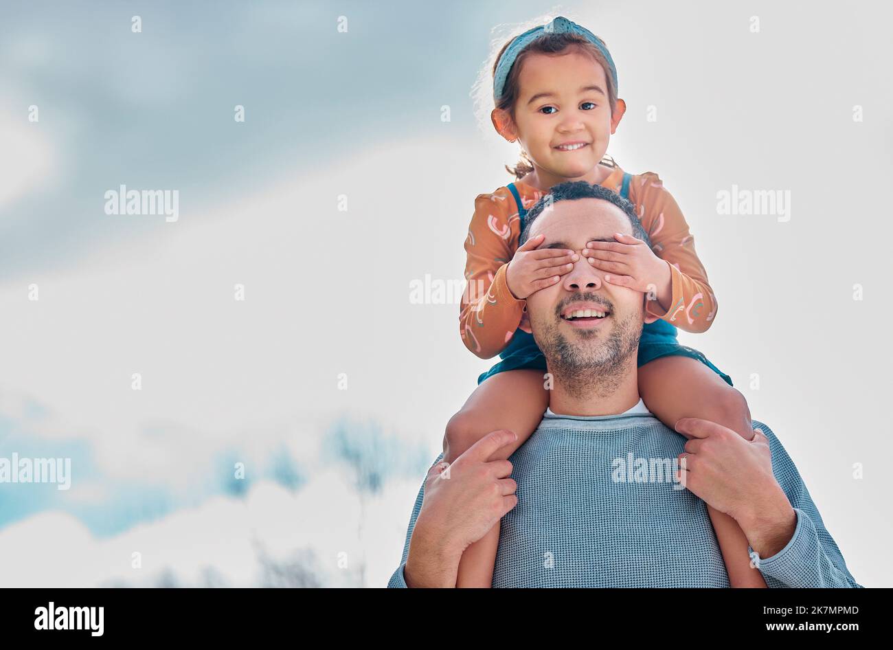 He doesnt know that its me. a little girl covering her fathers eyes while sitting on his shoulders outdoors. Stock Photo