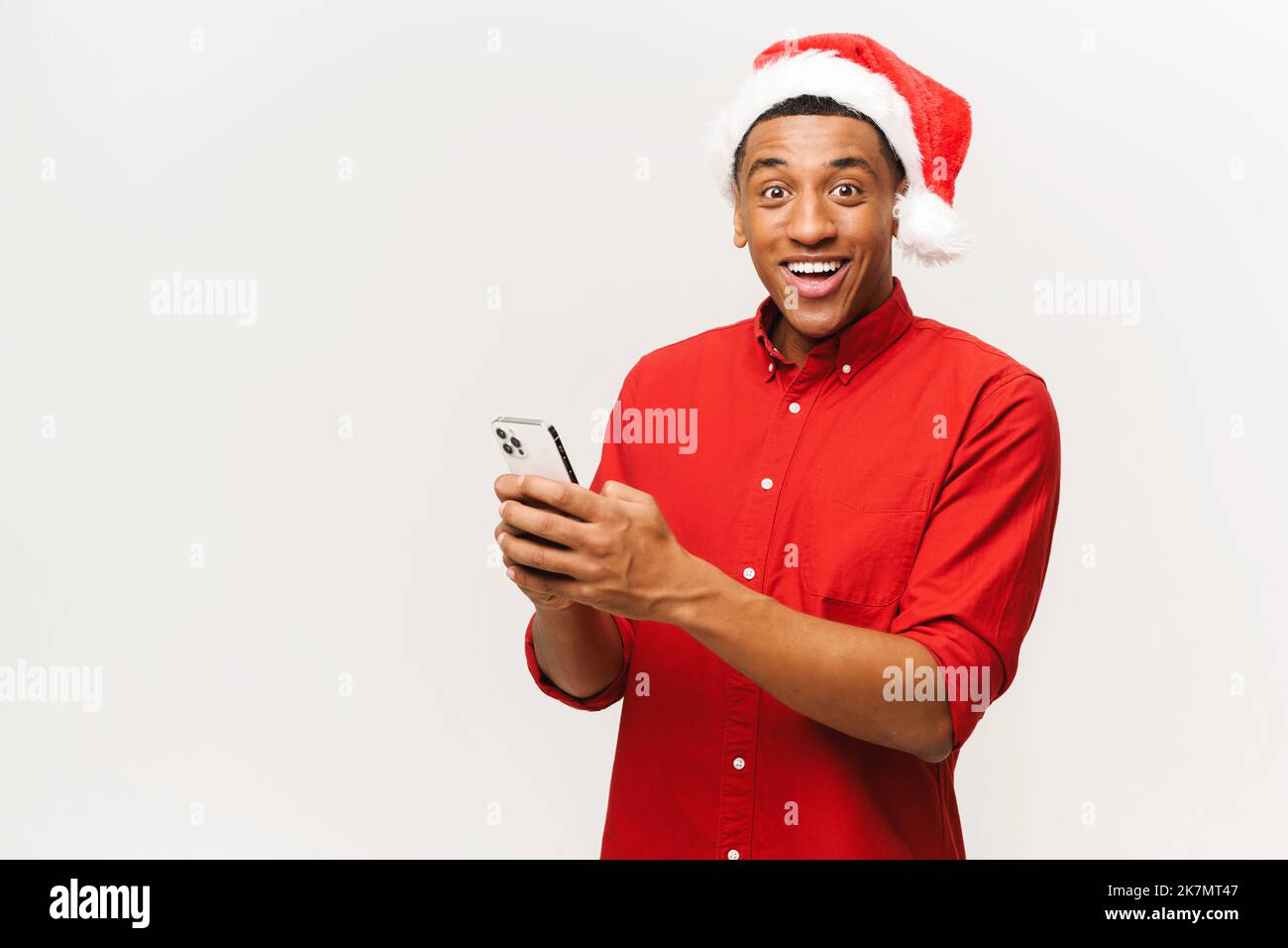 Young handsome happy shocked man in santa hat holds smartphone and looking at the camera with surprised opened mouth, confused cheerful male, isolated on white. New Year advertising concept Stock Photo