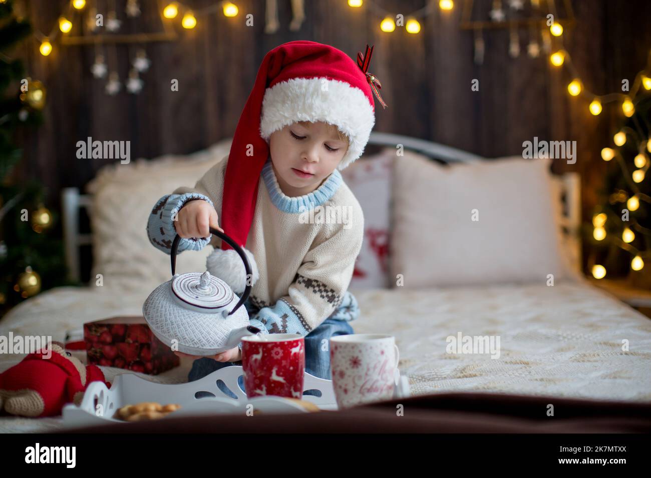 Cute toddler child, boy in a Christmas outfit, playing in a wooden cabin on Christmas, decoration around him. Child reading book and drinking tea Stock Photo