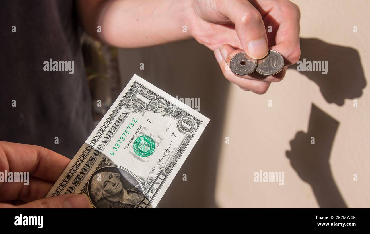 An Asian man hands over a US dollar bill in exchange for 150 JPY, which is the current approximate rate in October 2022. Their shadow reflect on the w Stock Photo