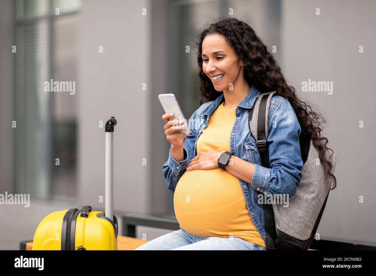 Cheerful young pregnant woman travelling alone, using smartphone at airport Stock Photo