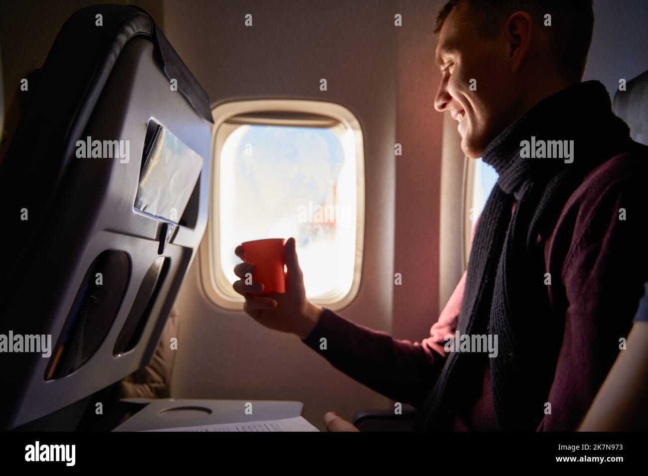 Smiling airline passenger with a paper cup of coffee looking ahead in airplane window. Man drinks tea onboard. Male holds beverage or water during Stock Photo