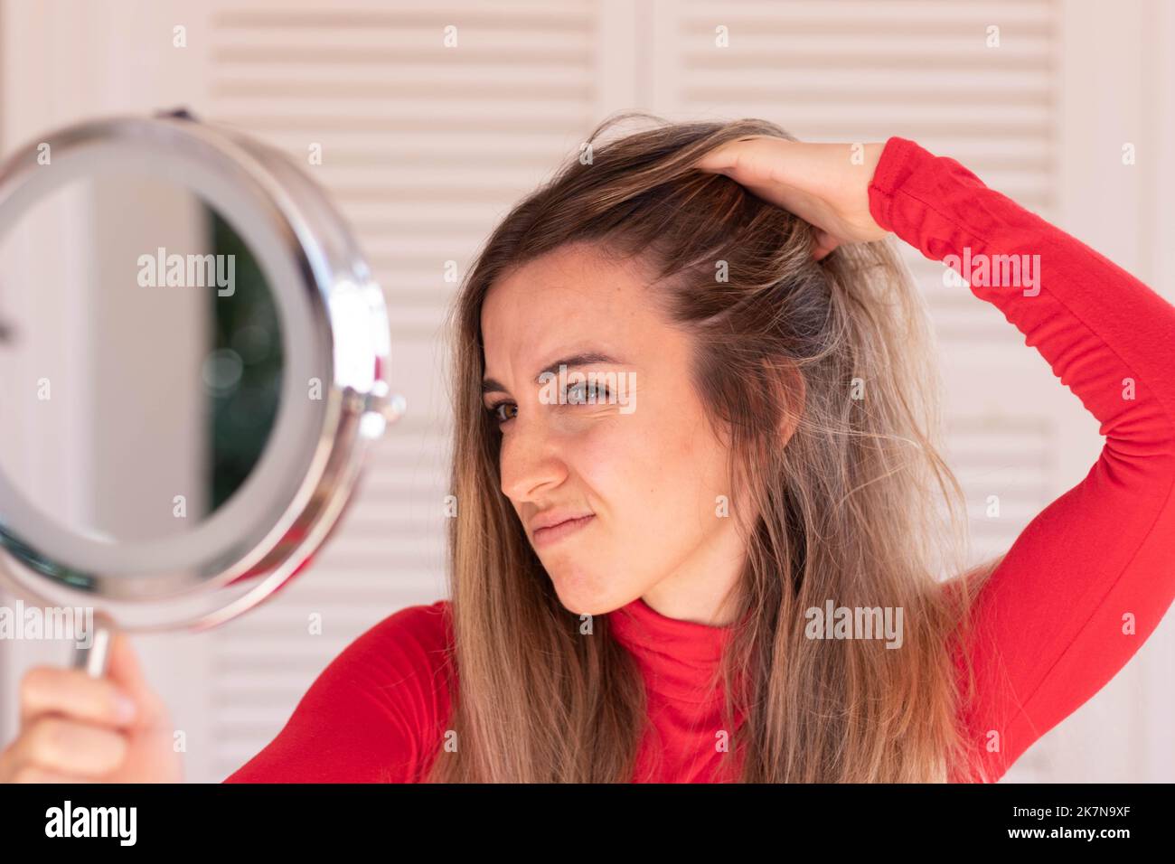 Young woman concern about her hair loss in autumn time while looking to round hand mirror in red outfit and white background Stock Photo