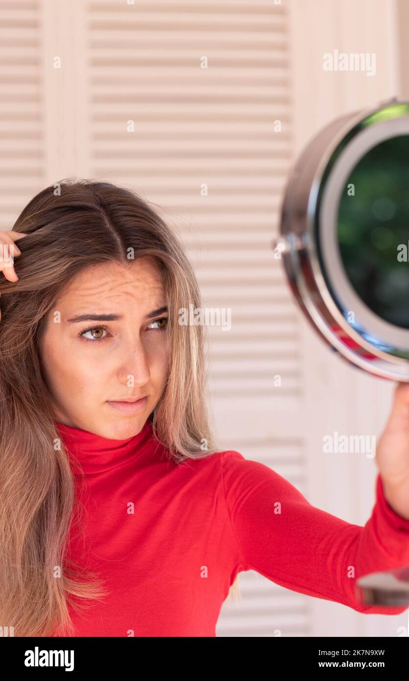 Vertical view of young blond woman looking the hair line in a round mirror about the hair loss in fall time in red outfit Stock Photo