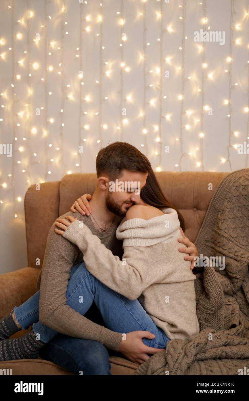 Happy married young couple hugging, sitting on cozy couch together, woman and man enjoying leisure time, relaxing on sofa in living room at home with lights in background Stock Photo