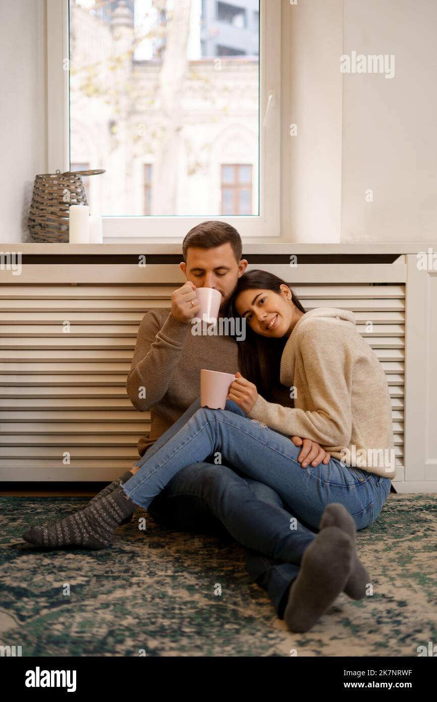 Romantic couple at home. An Attractive young woman and handsome man are enjoying spending time together while siting coddling with cups of tea in hands. Stock Photo