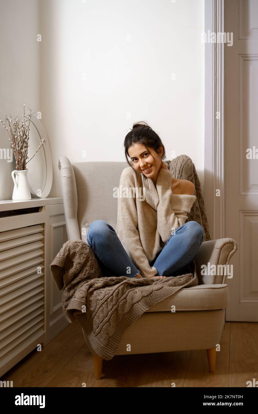 Smiling lady in smart trendy wear is sitting on armchair. Smiling, sitting in relaxing atmosphere indoors at home, nice design. Comfort and cozy girlish time Stock Photo