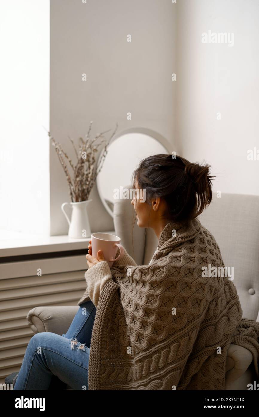 Smiling lady in smart trendy wear is sitting on armchair with a cup of tea. Smiling, sitting in relaxing atmosphere indoors at home, nice design. Stock Photo