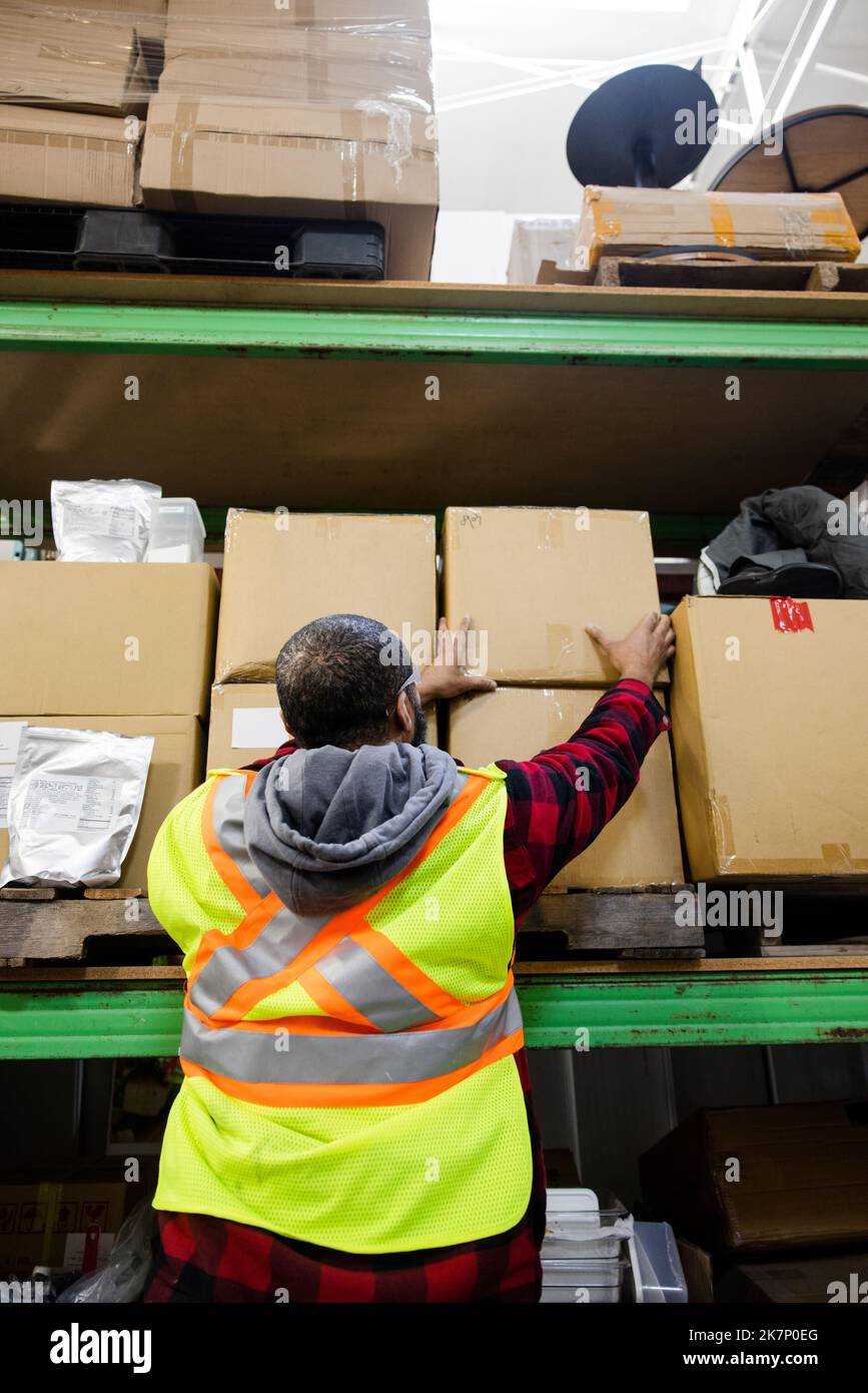 Male warehouse worker in reflective vest reaching for box on shelf