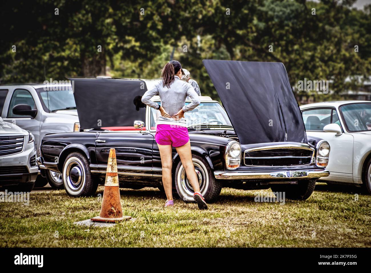 Antique car show. Woman in pink shorts checks out vintage car with hood and truck up surrounded by various other cars in grassy area with old traffic Stock Photo