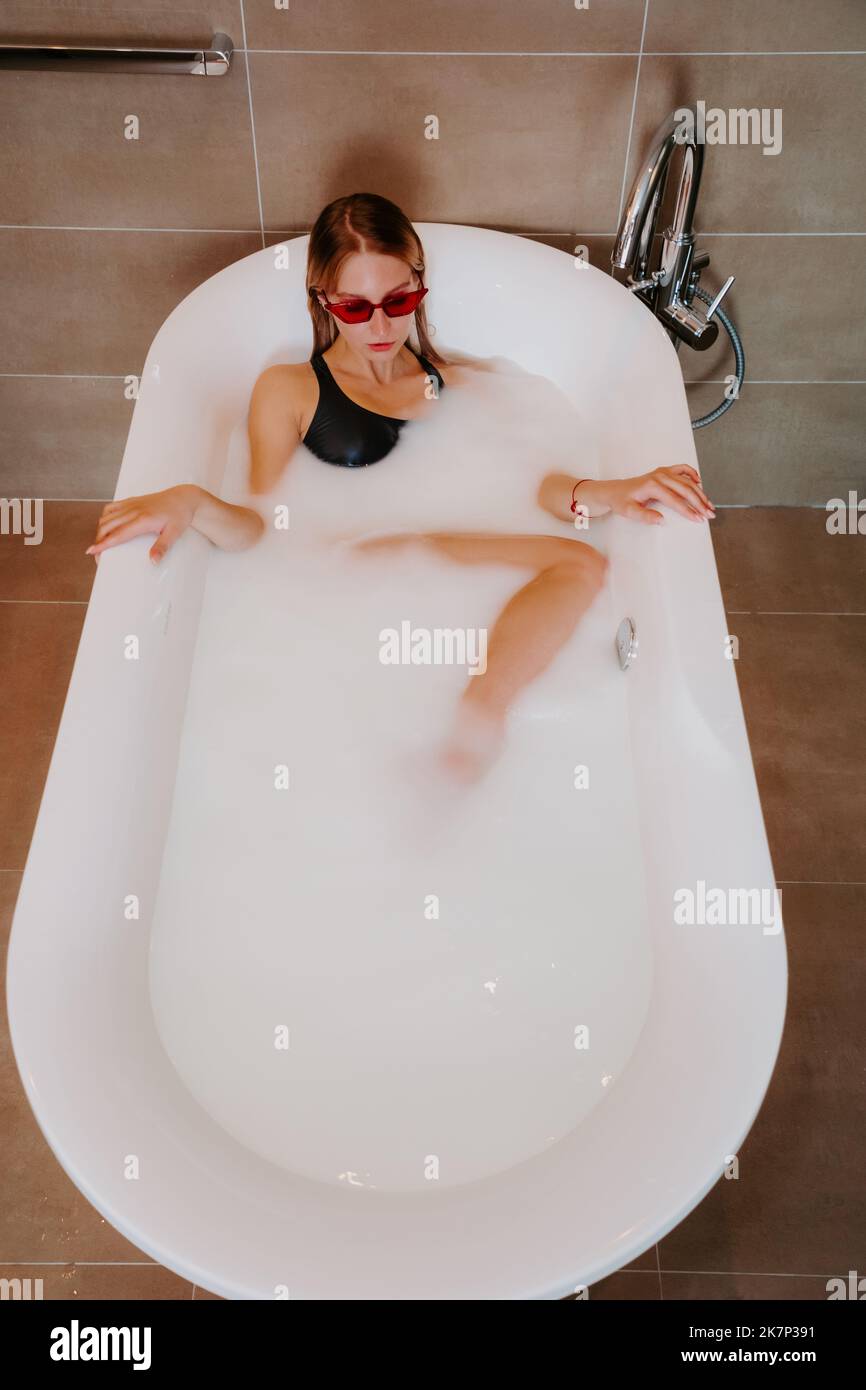 Pretty blonde female, wearing a black swimsuit, in white bathtub filled with milk Stock Photo