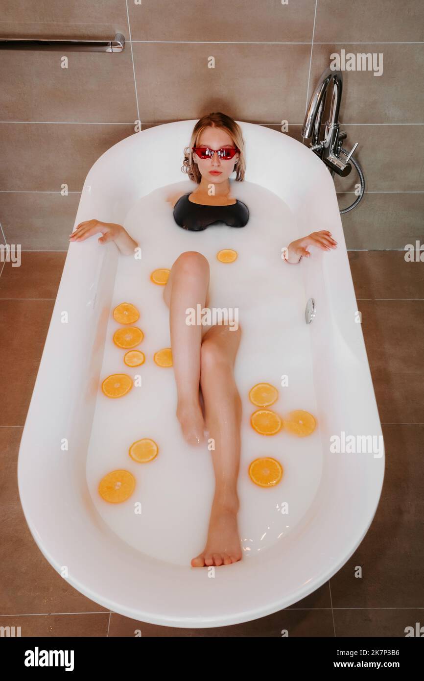 Pretty blonde female, wearing a black swimsuit and red sunglasses, in white bathtub filled with milk and oranges Stock Photo