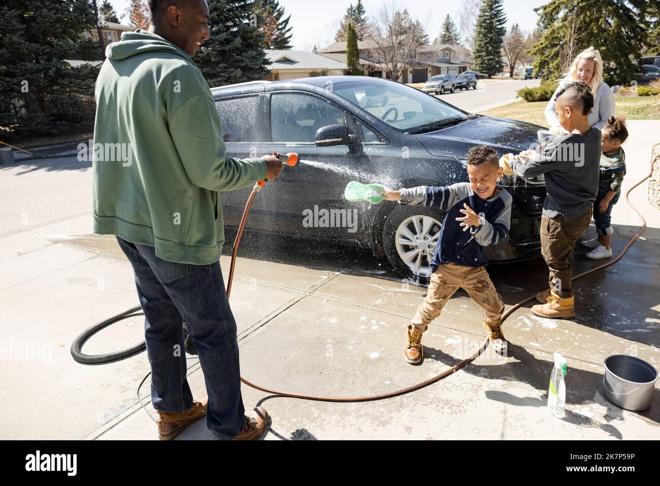 Father spraying water at son, family washing car in driveway Stock