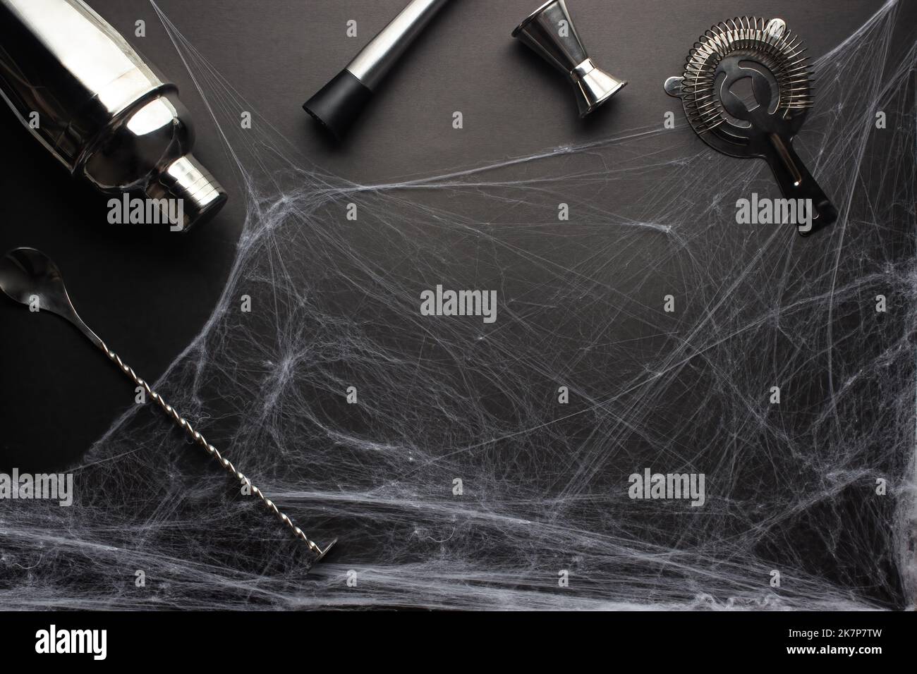 Various bar accessories such as: a shaker, muddler, strainer, as well as a bar spoon and a measuring stack lie against the backdrop of a spider web on Stock Photo