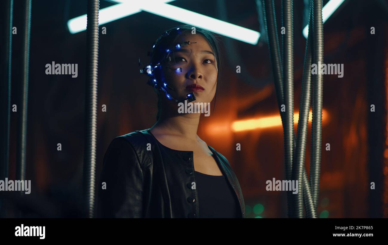 Cyberpunk girl in a black leather jacket and intense facial expressions. Asian girl with one-eyed glasses and headset. Trapped behind black hanging rods. Cyborg, sci-fi, Neon lights. Stock Photo