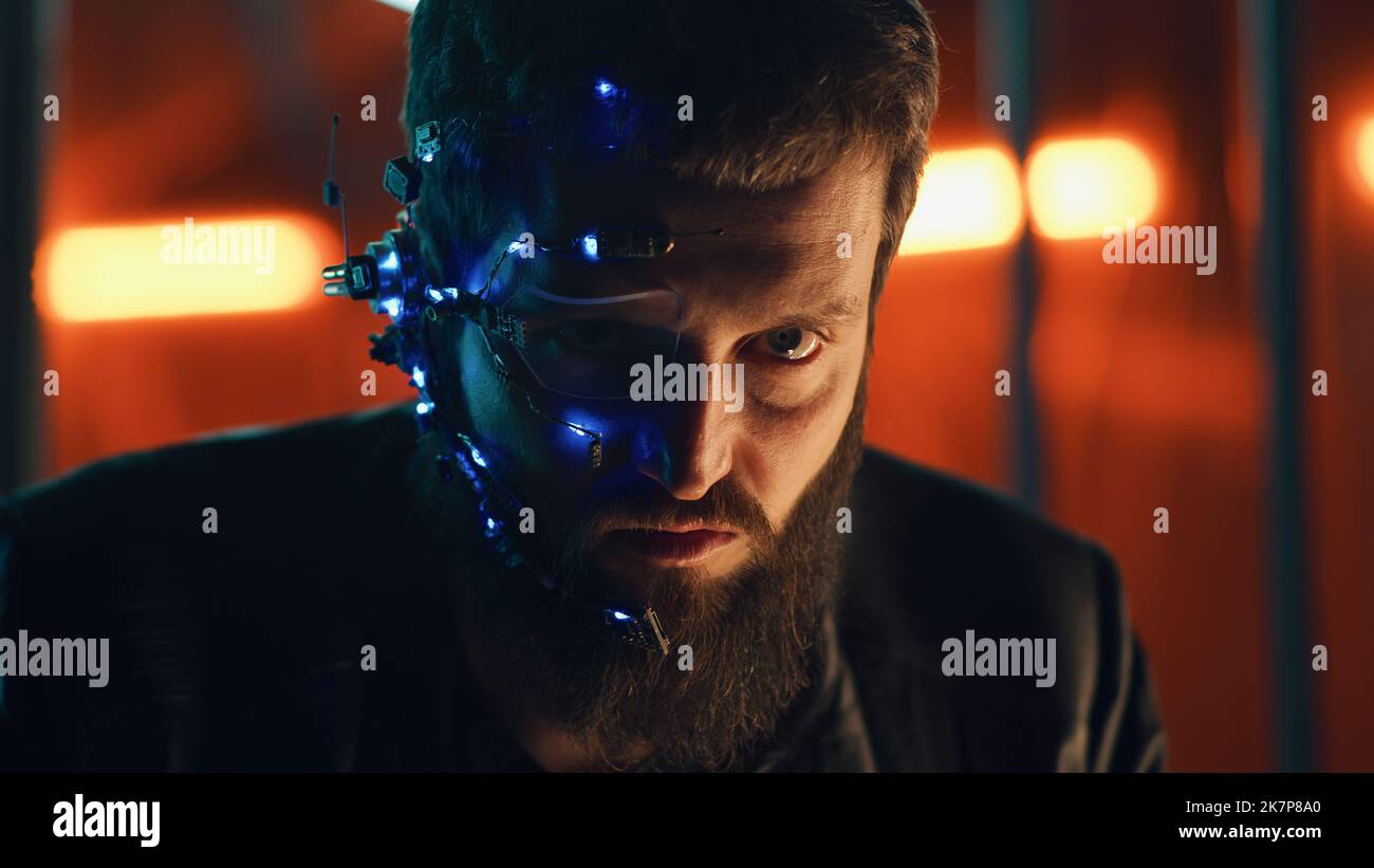 Brunette guy with dark beard wearing futuristic headset with one-eyed glasses, mic and ear piece looks at the camera with intense facial expressions. Neon lights in the background. Cyberpunk. Stock Photo