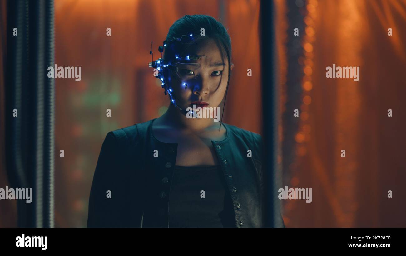 Cyberpunk girl in a black leather jacket trapped behind black hanging rods. Intense facial expressions. Asian girl with futuristic glasses and headset. Cyborg, sci-fi, Neon lights. Stock Photo