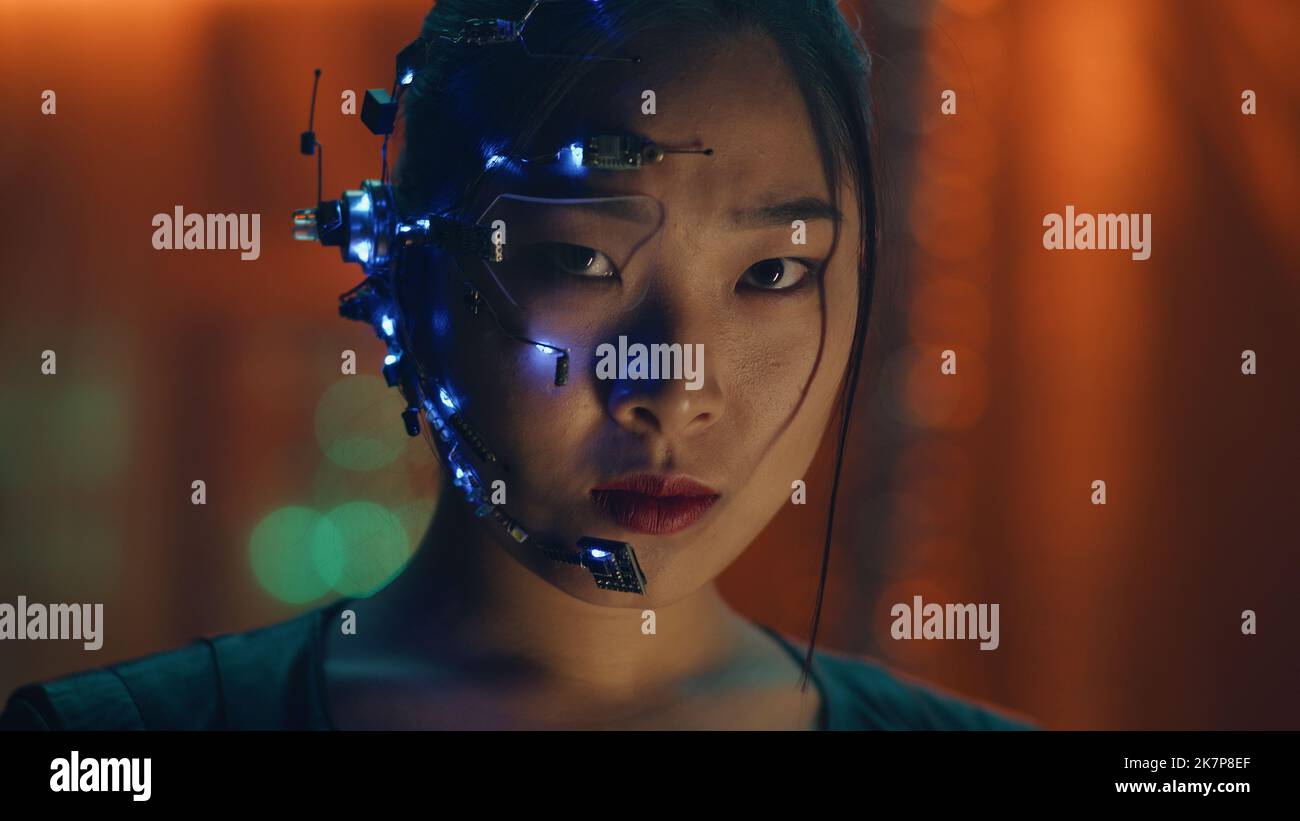 Asian girl in Cyberpunk style with a head set and microphone with small white LED lights looks at the camera. Intense facial expressions. Sci-fi background with Neon lights. Stock Photo