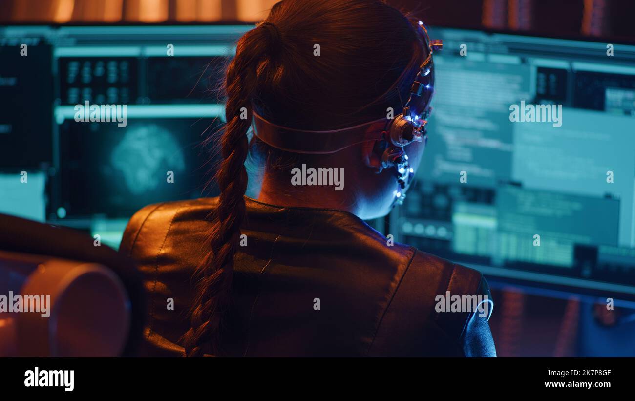 Back view of a Cyberpunk girl types on the keyboard. Multiple computer screen with strings of codes infront of her. Devloping codes for futuristic software. Neon lights in the background. Stock Photo