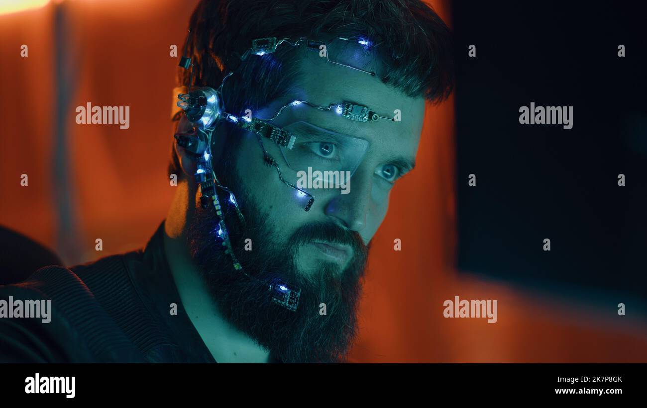 Brunette guy works on the computer with neon lights in background. Cyberpunk. Wearing a futuristic one-eyed glasses and microphone. Cyber and sci-fi backgrounds. Stock Photo