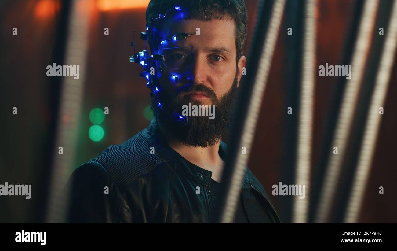 Young guy wearing a headset with LED lights stands behind the black hanging rods and looks at the camera. Cyberpunk and futuristic concept. Neon lights in the background. Stock Photo