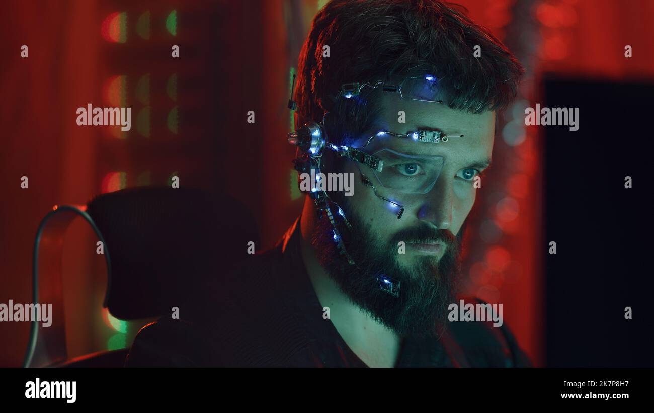 A Cyberpunk guy looks at the computer screen. Wearing futuristic one-eyed glasses with earpiece and microphone. Cyber and sci-fi backgrounds. Stock Photo