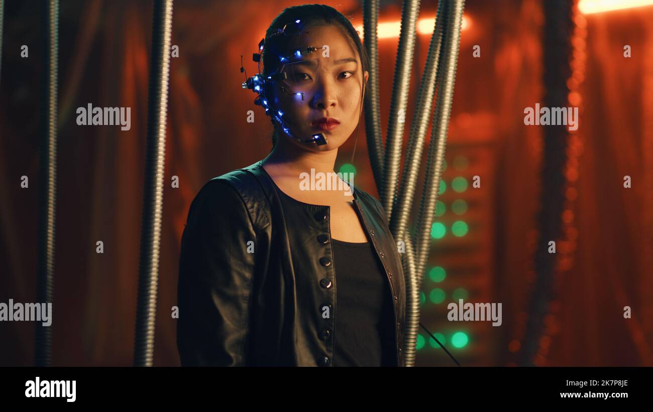 Cyberpunk girl in a black leather jacket looks fiercely at the camera. Asian girl with one-eyed glasses and headset trapped. Black thick rods hanging to prevent her from escaping. Stock Photo