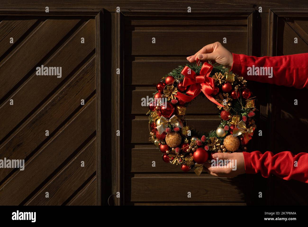 Female hands decorate the wall of a wooden house with a festive Christmas wreath. The concept of decorating the facade of the house and preparing for Stock Photo