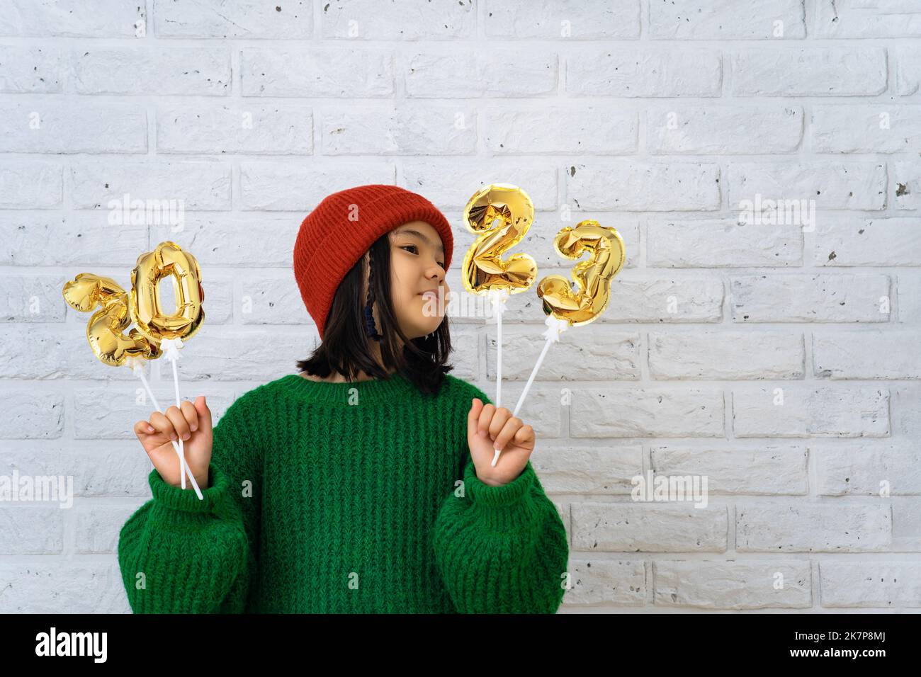 Asian cute girl in red cap and green sweater holding foil balloons with numbers 2023. Copy space, brick white wall background, face turned to the side Stock Photo