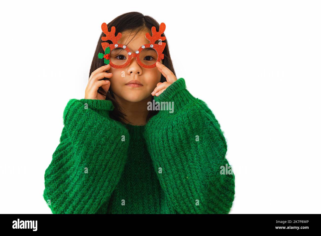 Beautiful Asian child girl in a green sweater puts on masquerade Christmas glasses with deer horns. Portrait Beautiful Vietnamese girl wearing Christm Stock Photo