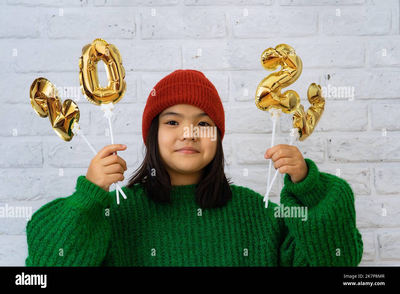 Asian child girl in a hat and a green sweater close-up, in the hands of golden balloons with the numbers 2023, copy space, against a white brick wall, Stock Photo