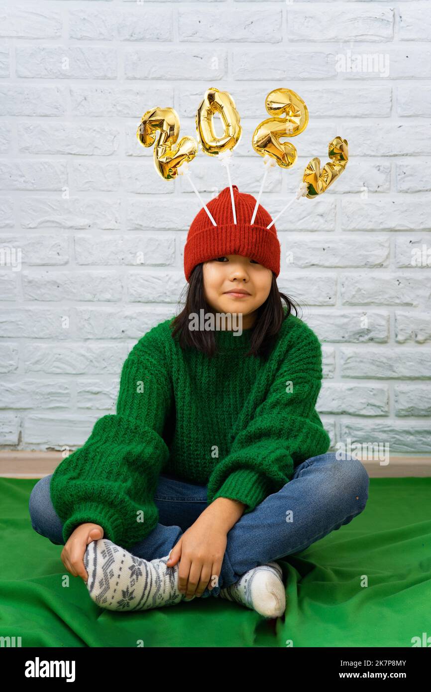 Funny Asian girl in a green sweater sits on the floor with golden balloons with the number 2023 in a red cap. A girl sits cross-legged on the floor wi Stock Photo