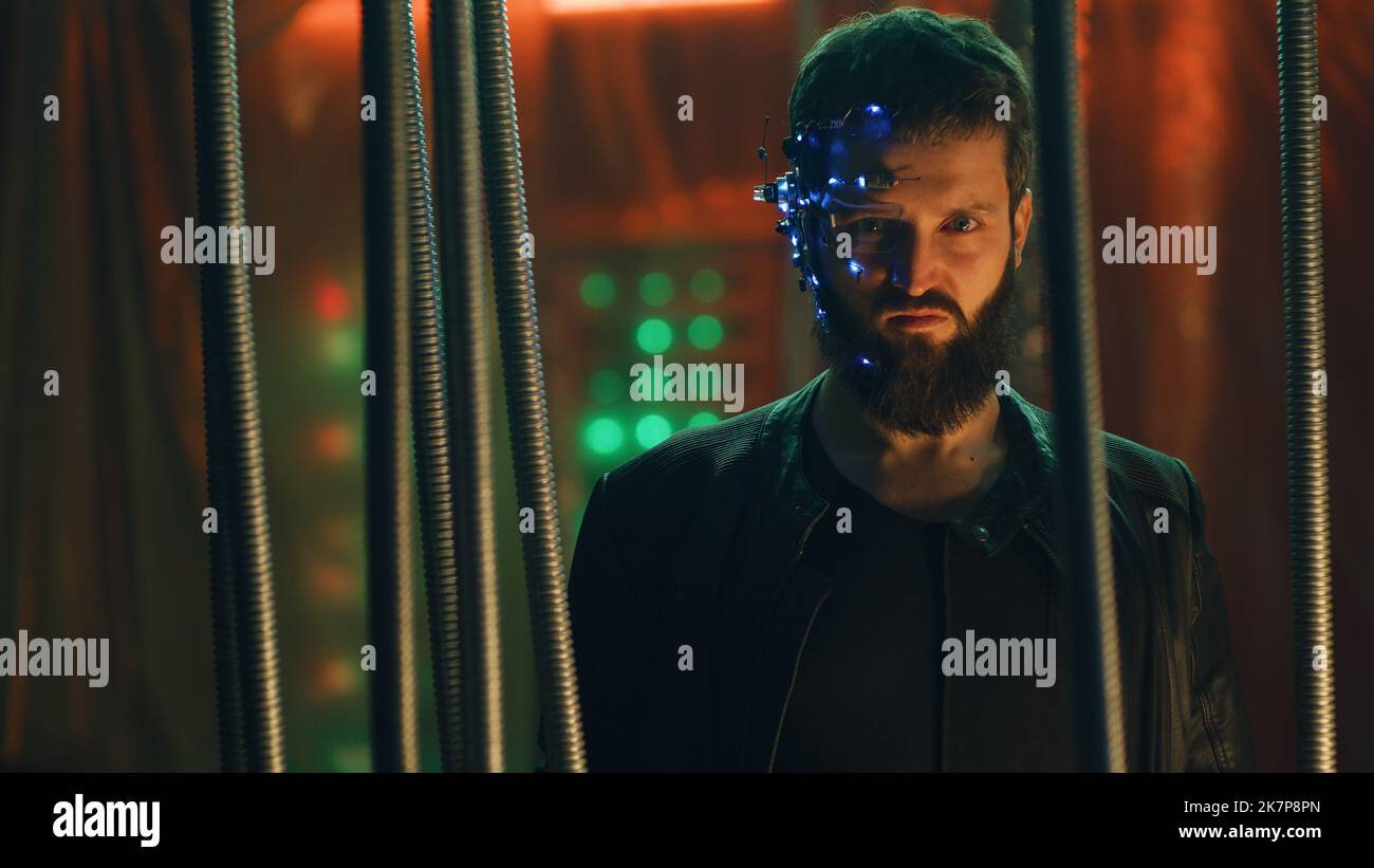 Young brunette guy in Cyberpunk style stands behind the black rods hanging. Wearing a futuristic headset with eyepiece and microphone. Neon lights background. Science fiction. Stock Photo