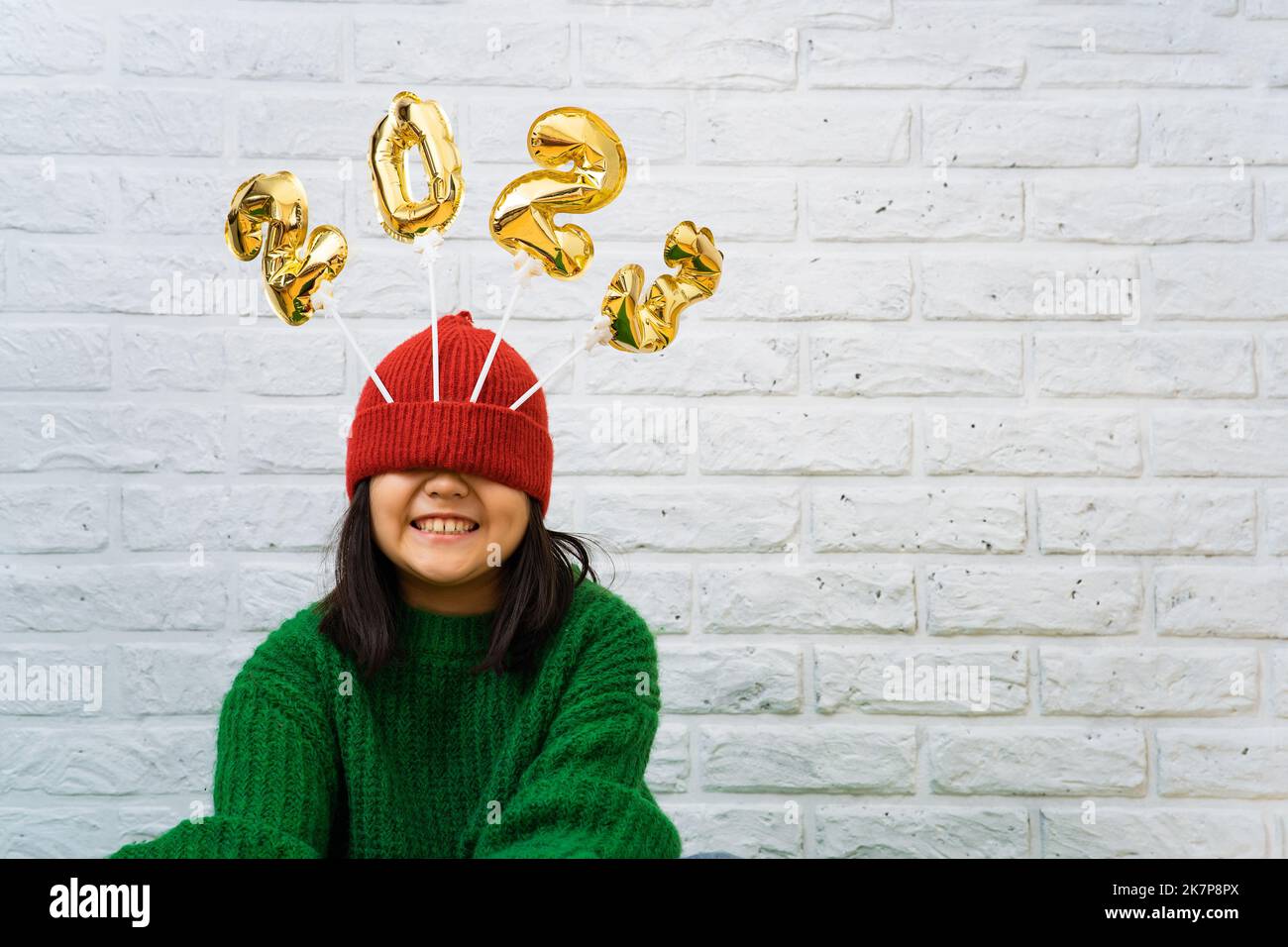 Happy Asian baby girl in a green sweater close-up with golden balloons with numbers 2023 in hat. Copy space, background of a white brick wall, the eye Stock Photo