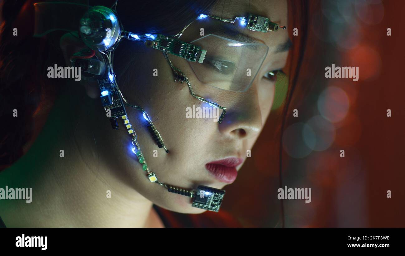 Focused asian girl in cyberpunk attire works on the computer. Wearing one-eyed glasses with white LED lights and a microphone. Neon lights background. science fiction, cybernetics concept. Stock Photo