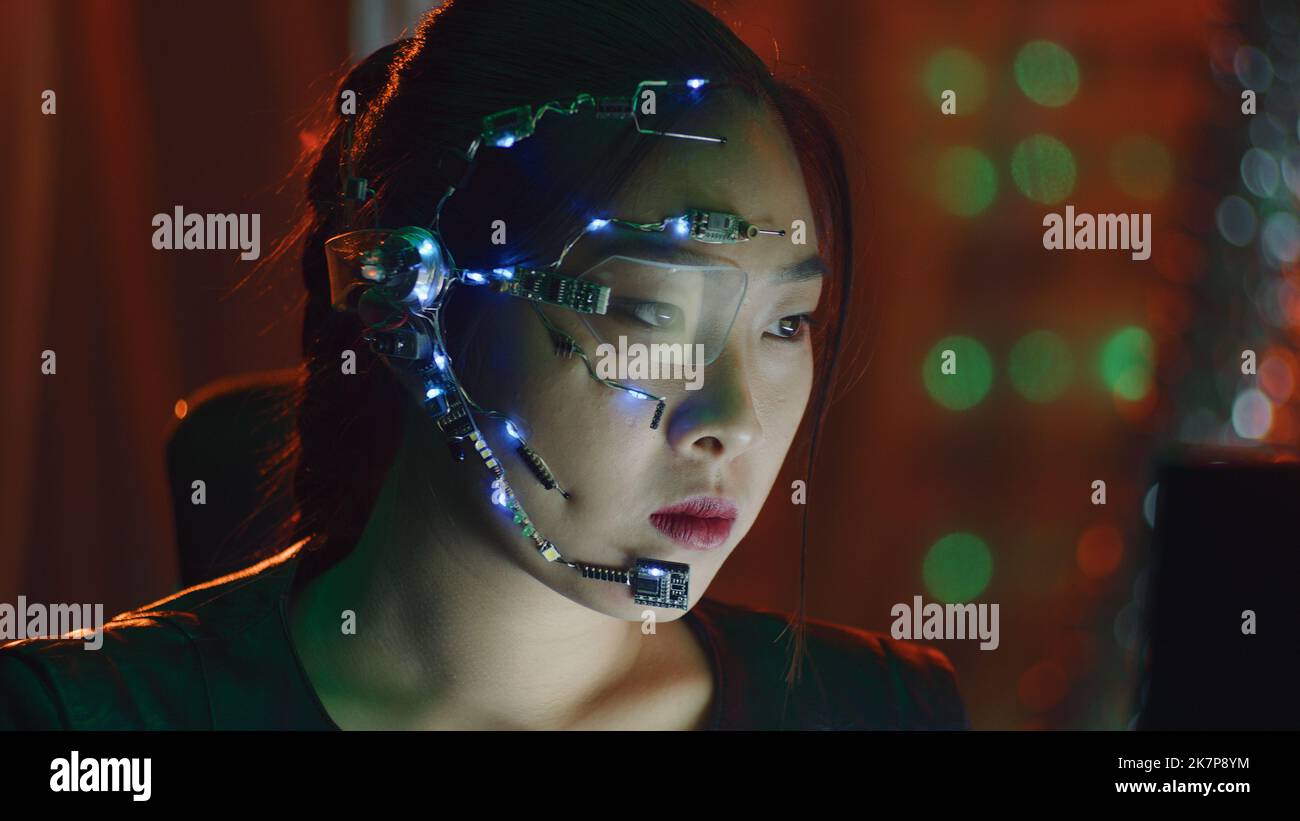 A Cyberpunk girl works on the computer in the red neon lights. Asian girl with futuristic one-eyed glasses and microphone. Cyber and sci-fi backgrounds. Stock Photo