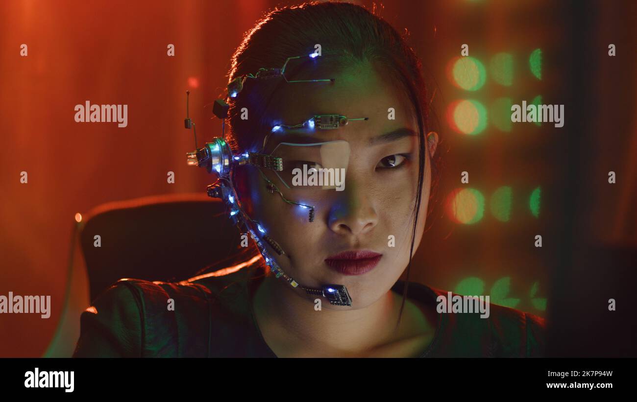 A Cyberpunk girl looks at the computer screen. Wearing futuristic one-eyed glasses with microphone. Stops looking at the computer to face the camera. Cyber and sci-fi backgrounds. Stock Photo