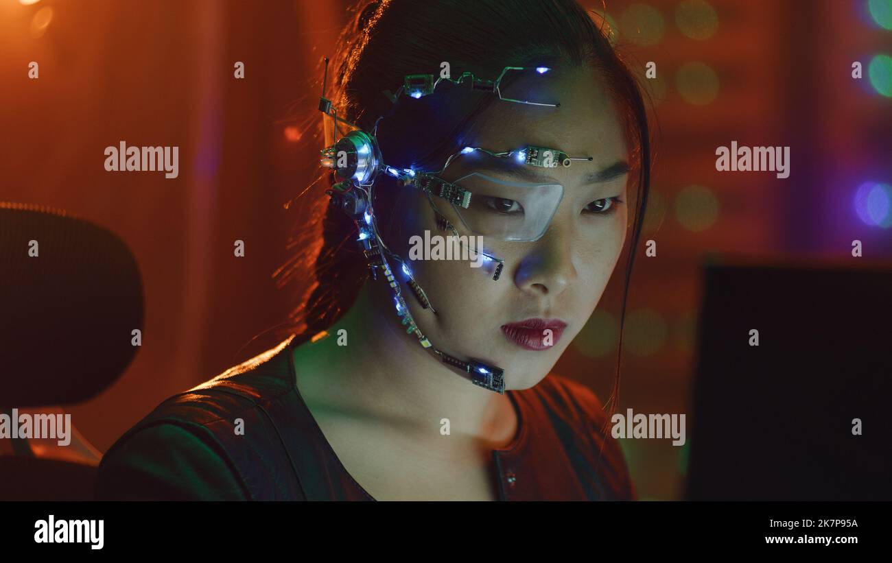 A Cyberpunk girl works on the computer in the red neon lights. Asian girl with futuristic one-eyed glasses and microphone. Cyber and sci-fi backgrounds. Stock Photo