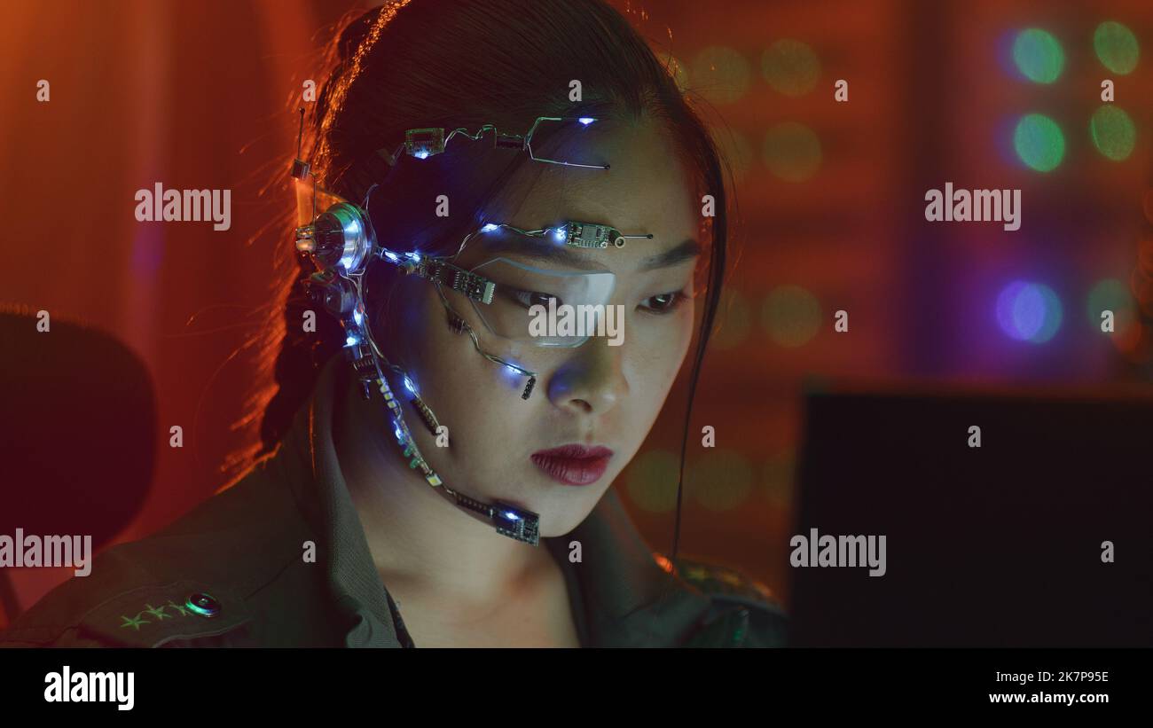 Focused asian girl in cyberpunk attire works on the computer. Wearing one-eyed glasses with white LED lights and a microphone. Neon lights background. science fiction, cybernetics concept. Stock Photo