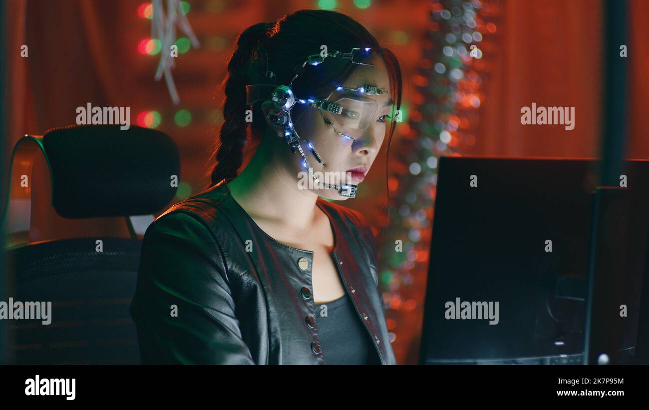 Focused cyberpunk girl in black leather jacket works on the computer. Wearing a headset with white LED lights and microphone. Cyberpunk background with neon lights. Stock Photo