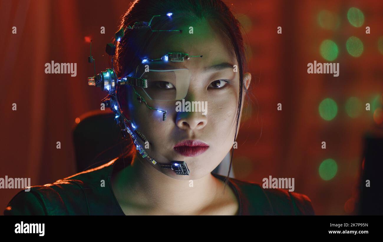 Asian girl in cyberpunk style wearing one-eyed glasses and microphone with small LED lights looks at the camera with serious facial expression. Sci-fi background with neon lights. Stock Photo