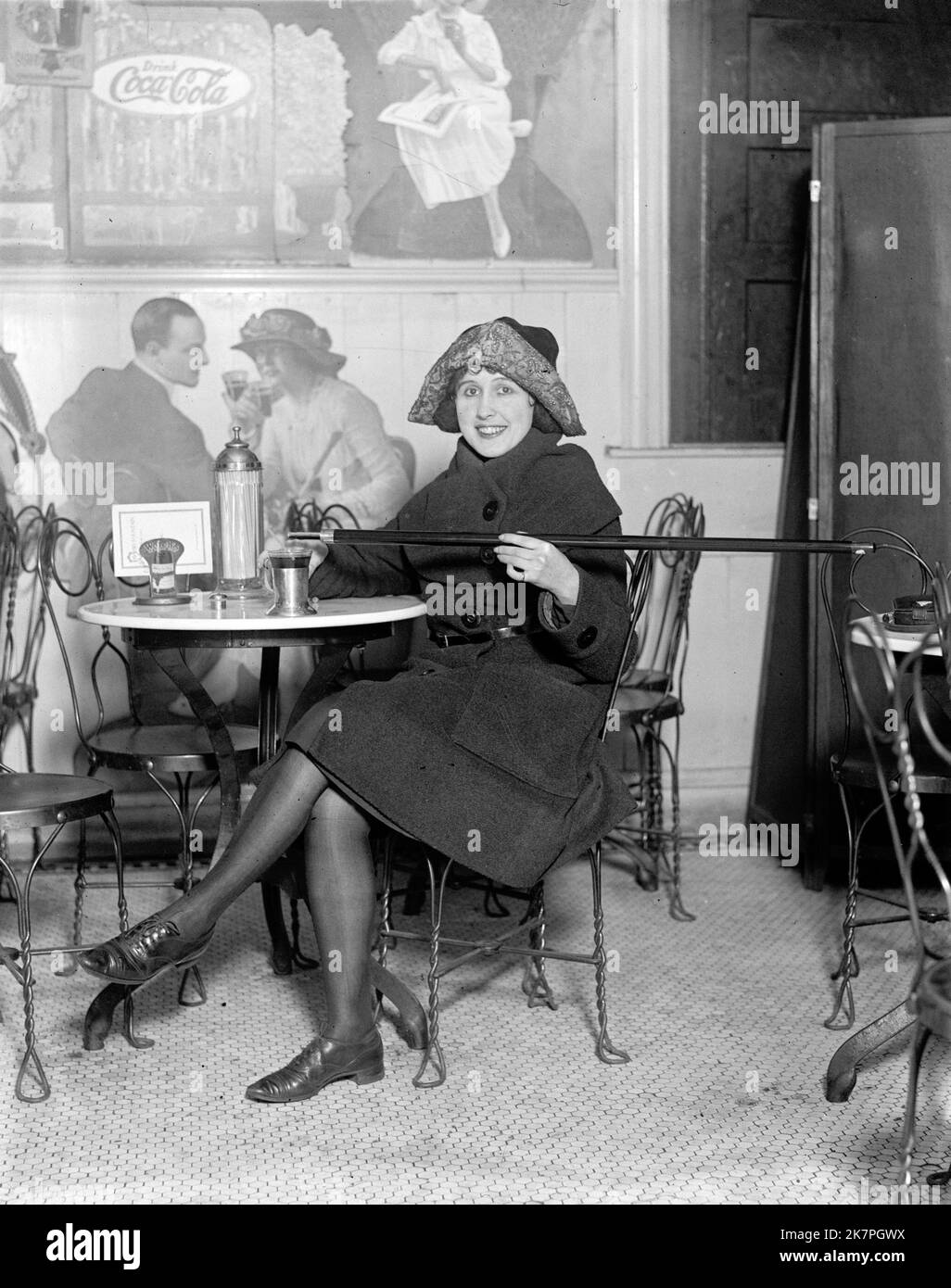 Woman seated at a soda fountain table is pouring alcohol into a cup from a cane, during Prohibition; with a large Coca-Cola advertisement on the wall, America Stock Photo