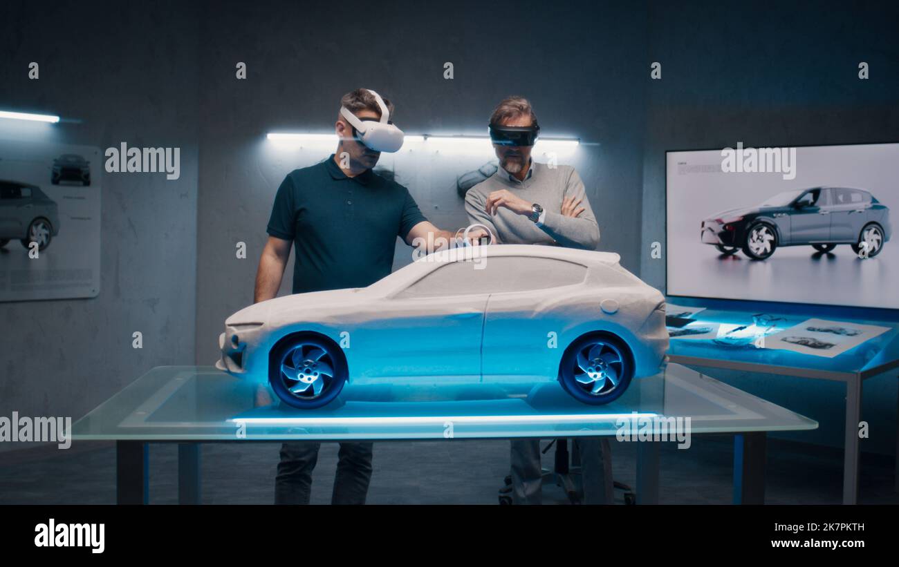 Automotive professional engineers with VR headset and joysticks talk about vehicle production while standing infront of a prototypecar model in a high tech office. Car design analysis and improvement. Stock Photo