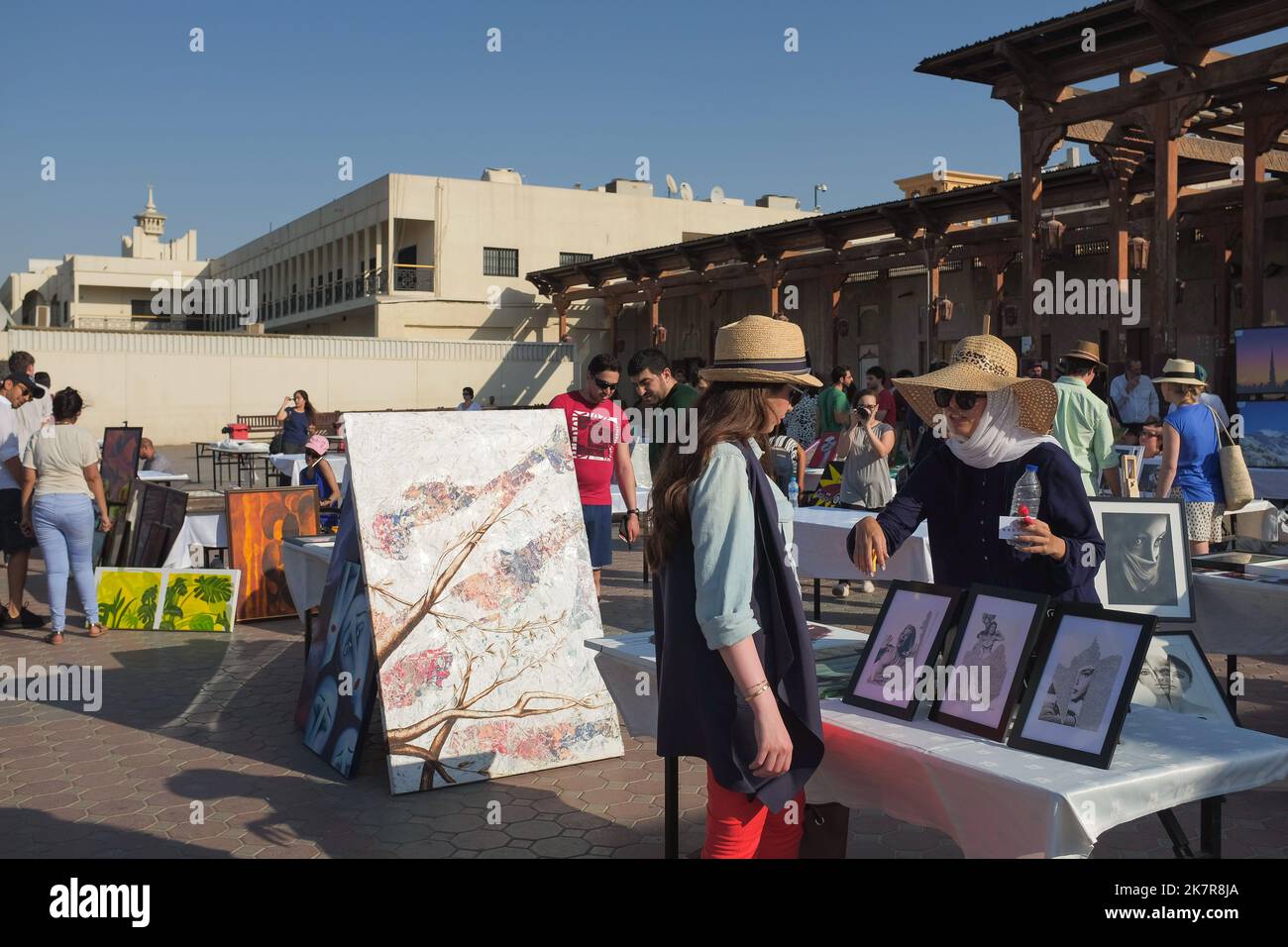 A woman visits a table with framed artworks at an outdoor art fair in historical Al Fahidi in Dubai, UAE. Female artist points and talks about her work. Stock Photo