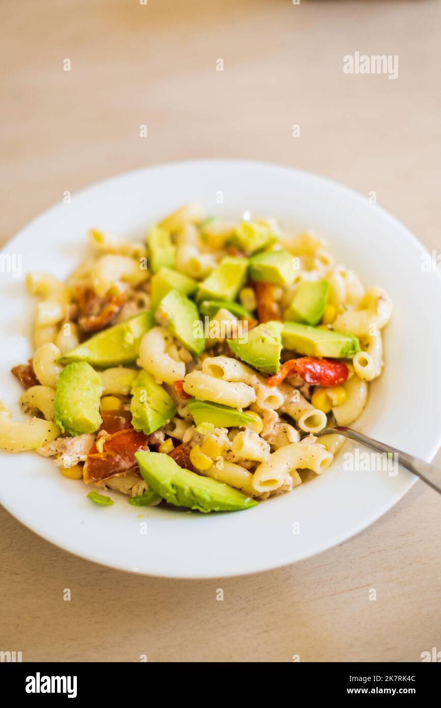 A cold pasta salad consisting of large elbow macaroni, slice avocados, toasted red bell peppers and corn in a white dish sat on a wood table. USA. Stock Photo